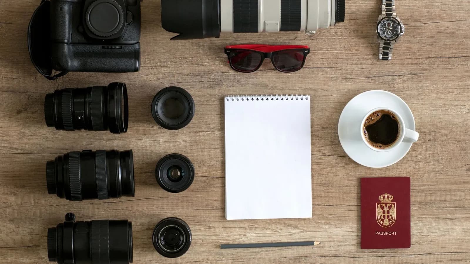 Matériel de voyage à plat : boîtier reflex, télézoom blanc, objectifs, carnet, café et passeport.