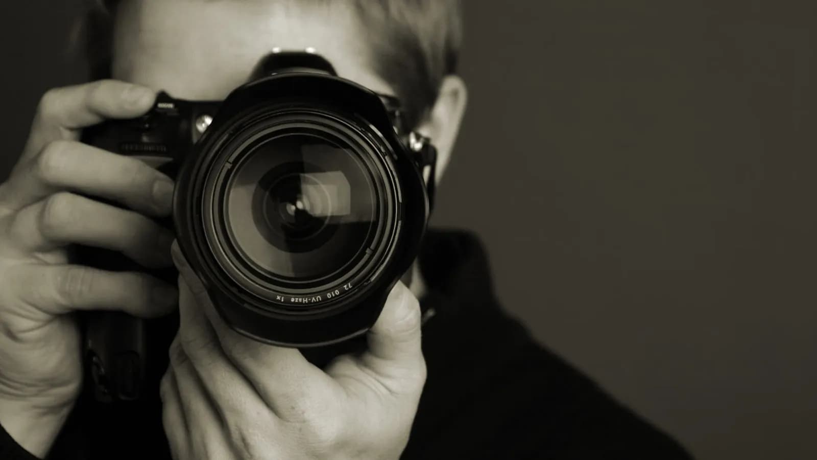 Gros plan d'un photographe tenant un reflex, objectif pointé vers l'objectif, arrière-plan flou, photo en noir et blanc.